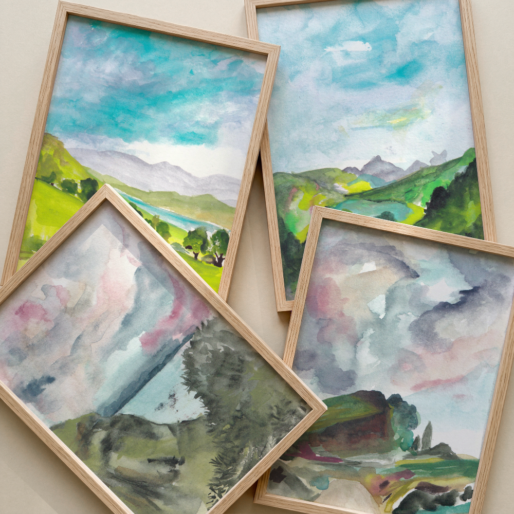 Four framed watercolor landscape paintings arranged overlapping on a light beige wall. The paintings depict green hills, blue skies, mountains, and soft pink and gray clouds, each in a light natural wood frame.